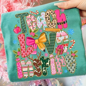 Pu&ograve; includere: T-shirt turchese con le parole "Teach Love Inspire" in un design colorato e fantasia. Le lettere sono decorate con vari elementi festivi e scolastici, tra cui mele, matite e fiocchi di neve. Una mano tiene la maglietta.