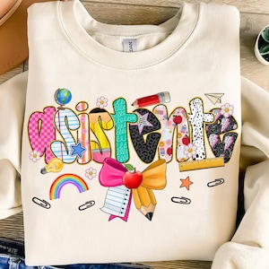 Può includere: Felpa color crema con la scritta "Assistant" in un font colorato e giocoso. Le lettere sono decorate con grafiche a tema scolastico come un mappamondo, una matita, una mela e un aeroplanino di carta. Un grande fiocco e un disegno di matita sono in basso.