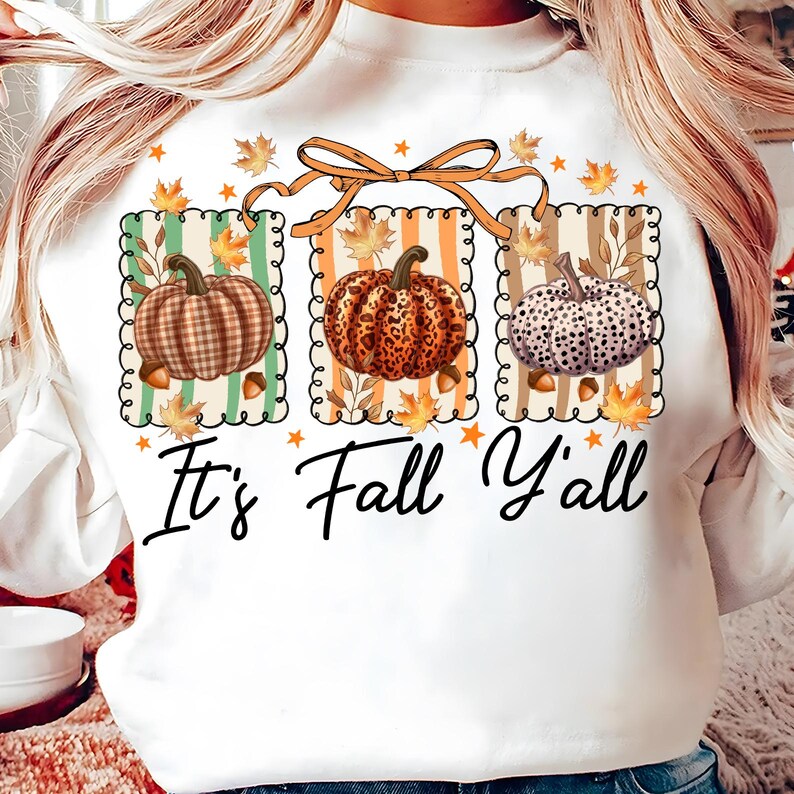 May include: White long-sleeve shirt featuring a fall-themed graphic. The design includes three pumpkins with different patterns, fall leaves, and the text "It's Fall Y'all". The pumpkins are set against striped backgrounds and are connected by an orange bow.