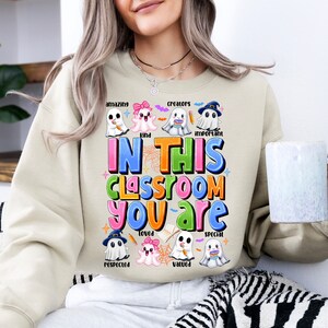 May include: A beige sweatshirt with a Halloween-themed design featuring ghosts and the text "In this classroom you are..." with words like "amazing", "kind", "creators", "important", "respected", "loved", "valued", and "special" written below each ghost.