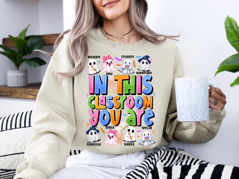 May include: A beige sweatshirt with a Halloween-themed design featuring ghosts and the text "In this classroom you are..." with words like "amazing", "kind", "creators", "important", "respected", "loved", "valued", and "special" written below each ghost.