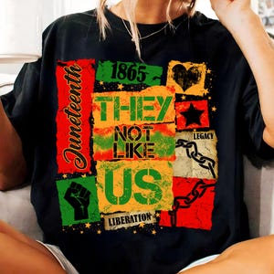 May include: Black t-shirt with a colorful graphic design celebrating Juneteenth. The design includes the text "1865," "THEY NOT LIKE US," and "LIBERATION," along with a fist, a heart, a star, and a chain. The word "Juneteenth" is written vertically.