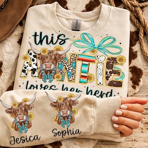 May include: Cream-colored sweatshirt with the text "this Auntie loves her herd." The word "Auntie" is decorated with a cow, sunflowers, and turquoise accents. The sleeve features two cow illustrations and the names "Jesica" and "Sophia."