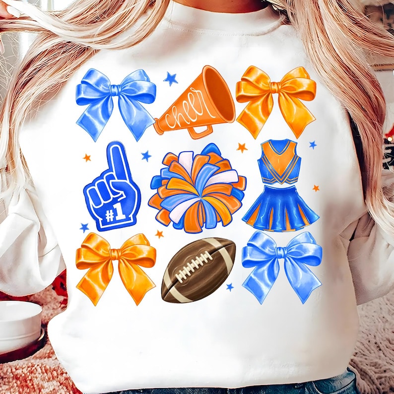 May include: White sweatshirt featuring a collection of cheerleading-themed graphics. Includes blue and orange bows, a megaphone with the word "cheer", a foam finger, a pom-pom, a cheerleader uniform, a football, and stars.