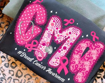 May include: A black t-shirt with the letters "CMM" in pink, featuring a stethoscope and pink ribbons. The letter "C" has white polka dots, while the "M" has hearts and stars. The text "#Breast Cancer Awareness" is also visible.