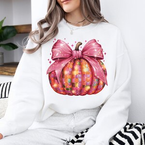 May include: White sweatshirt featuring a colorful pumpkin design. The pumpkin is decorated with a mosaic pattern and a large pink bow. The design is festive and suitable for fall or Halloween.