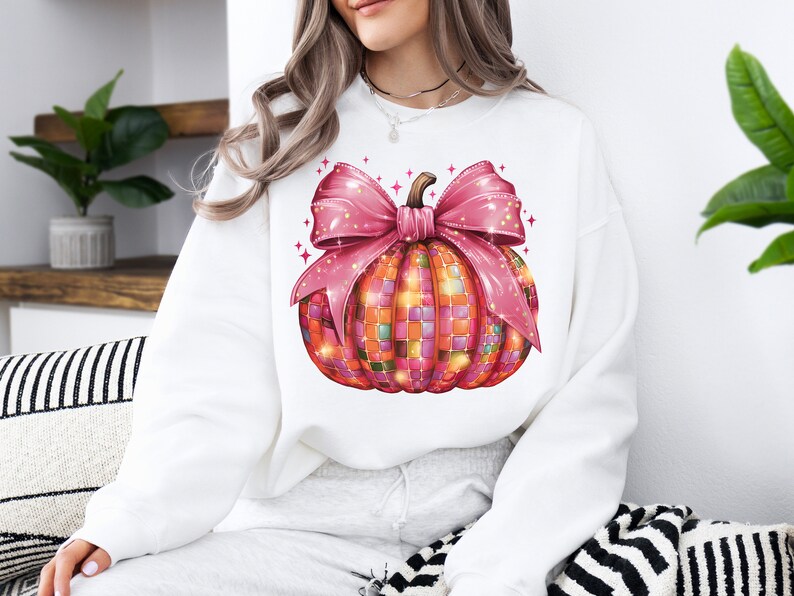 May include: White sweatshirt featuring a colorful pumpkin design. The pumpkin is decorated with a mosaic pattern and a large pink bow. The design is festive and suitable for fall or Halloween.