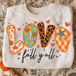 May include: Cream-coloured sweatshirt with the word "LOVE" in a fall-themed design. The "L" features pumpkins and rainbows, the "O" has autumn leaves, the "V" has a leopard print, and the "E" has a star. The phrase "fall y'all" is written below.