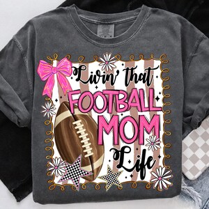 May include: Dark gray t-shirt with a graphic that says "Livin' that FOOTBALL MOM Life." The design includes a pink bow, a football, daisies, and stars. The text and design are in pink, white, and black.