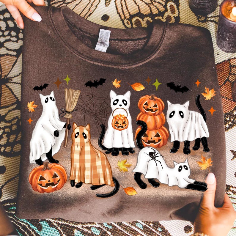 May include: A brown sweatshirt with a Halloween design. The design features cartoon cats dressed as ghosts, holding pumpkins, and wearing plaid. Bats, leaves, and pumpkins are also included in the design.