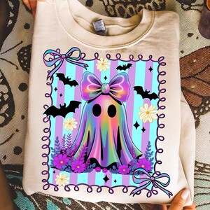 May include: Beige sweatshirt featuring a colorful ghost design. The ghost has a large bow and is surrounded by bats, flowers, and a striped background. The design includes purple, blue, and pink colors, perfect for Halloween.