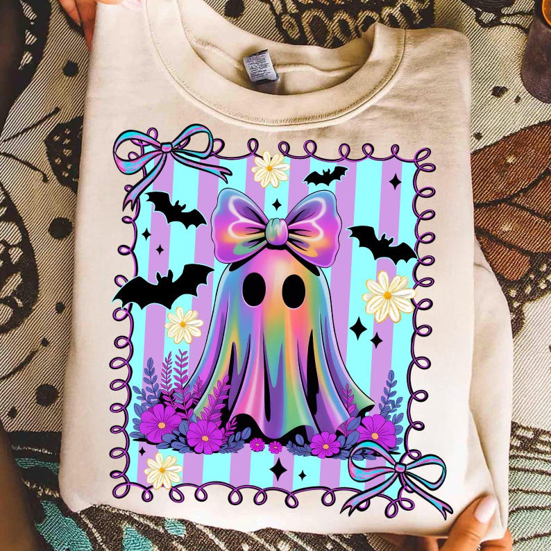 May include: Beige sweatshirt featuring a colorful ghost design. The ghost has a large bow and is surrounded by bats, flowers, and a striped background. The design includes purple, blue, and pink colors, perfect for Halloween.