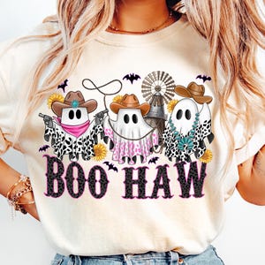 May include: Cream-colored t-shirt featuring a Halloween-themed graphic. The design includes three ghost figures wearing cowboy hats and bandanas, holding guns and a lasso. The text "BOO HAW" is printed in a black and pink font, with bats and sunflowers.