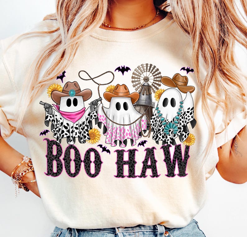 May include: Cream-colored t-shirt featuring a Halloween-themed graphic. The design includes three ghost figures wearing cowboy hats and bandanas, holding guns and a lasso. The text "BOO HAW" is printed in a black and pink font, with bats and sunflowers.