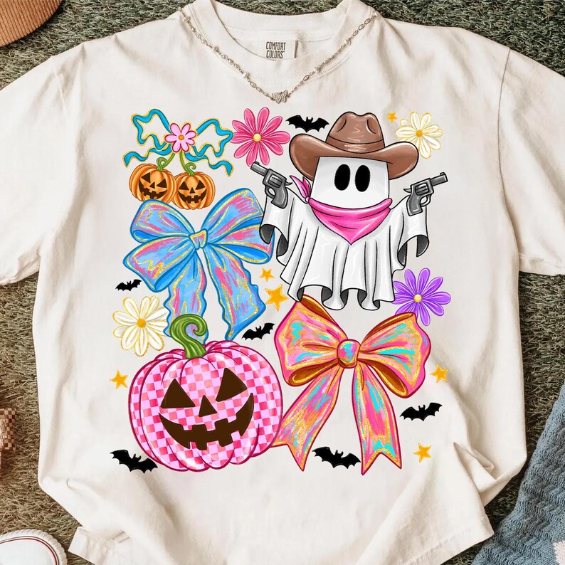 May include: Cream-coloured t-shirt with a Halloween graphic. The design features a ghost in a cowboy hat holding guns, alongside bows, pumpkins, flowers, and bats. The shirt is made of a soft material.