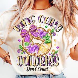 May include: A white t-shirt with a purple, green, and gold king cake design. The text "King Cake Calories Don't Count" is printed on the shirt.