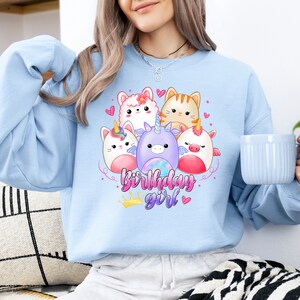 Puede incluir: Sudadera azul claro con un gr&aacute;fico de un unicornio, un gato y otros animales lindos. El texto "birthday girl" est&aacute; impreso debajo de los animales.