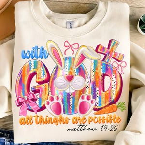 May include: A white sweatshirt with a colorful graphic of a bunny rabbit inside a rainbow striped egg that spells out "with God all things are possible Matthew 19:26".