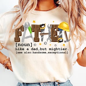 May include: Cream-colored t-shirt with the word "FATHER" in a brown, block letter design, incorporating tools like a wrench, saw, and hammer. The text below reads "Like a dad, but mightier."