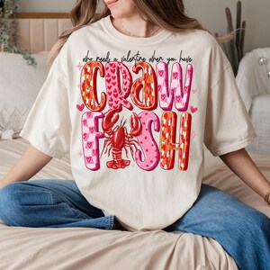 May include: A white t-shirt with a pink and red graphic that says "Who needs a valentine when you have crawfish".