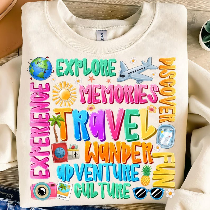 May include: A white sweatshirt with a colorful graphic design featuring the words "Explore", "Memories", "Travel", "Wander", "Adventure", "Culture", "Discover", "Fun", and "Experience". The graphic also includes a globe, airplane, sun, palm trees, pineapple, sunglasses, and a camera.