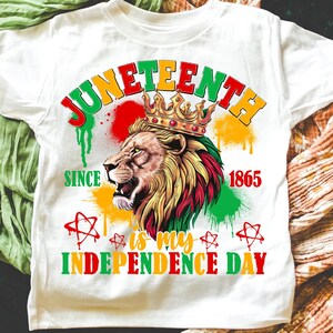 May include: White t-shirt featuring a lion wearing a crown, with text reading "Juneteenth" in red, yellow, and green. The shirt also includes the phrases "Since 1865" and "is my Independence Day."