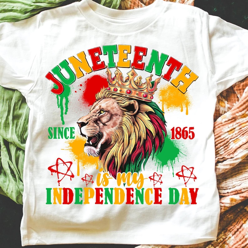 May include: White t-shirt featuring a lion wearing a crown, with text reading "Juneteenth" in red, yellow, and green. The shirt also includes the phrases "Since 1865" and "is my Independence Day."