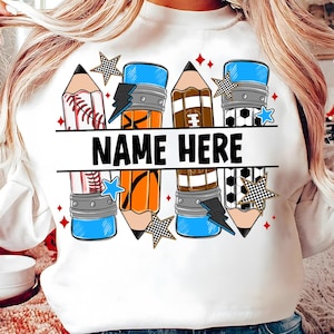 Pode incluir: Camisola branca com um design de l&aacute;pis com temas desportivos. O design inclui elementos de basebol, basquetebol e futebol, juntamente com o texto "NAME HERE" em preto. Acentuado com estrelas e raios.