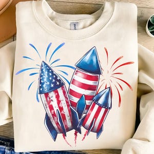 May include: Cream-colored sweatshirt featuring a graphic of three stylized rockets with red, white, and blue designs. The rockets are depicted with the American flag and surrounded by red and blue fireworks.