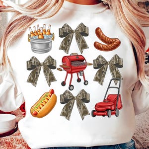May include: White sweatshirt featuring illustrations of a red grill, hot dog, sausages, beer in a bucket, a lawnmower, and camouflage bows. The design evokes a summer barbecue theme.