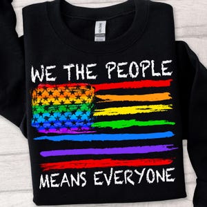 May include: Black sweatshirt with the text "WE THE PEOPLE MEANS EVERYONE" in white. The design features a rainbow-colored flag with stars. The sweatshirt is a Gildan Heavy Blend.