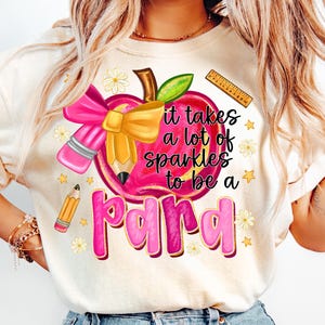 May include: Cream-colored t-shirt featuring a graphic design with a pink apple, pencil, and bow. The text reads "it takes a lot of sparkles to be a para." Additional elements include a ruler, stars, and flowers.