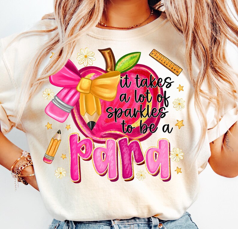 May include: Cream-colored t-shirt featuring a graphic design with a pink apple, pencil, and bow. The text reads "it takes a lot of sparkles to be a para." Additional elements include a ruler, stars, and flowers.