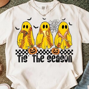 Puede incluir: Camiseta blanca con un dise&ntilde;o de Halloween. Tres figuras de fantasmas amarillos con costuras de b&eacute;isbol sostienen cada una un objeto de b&eacute;isbol. El texto "Tis' The Season" est&aacute; debajo, con calabazas talladas. Tambi&eacute;n hay murci&eacute;lagos y una telara&ntilde;a.