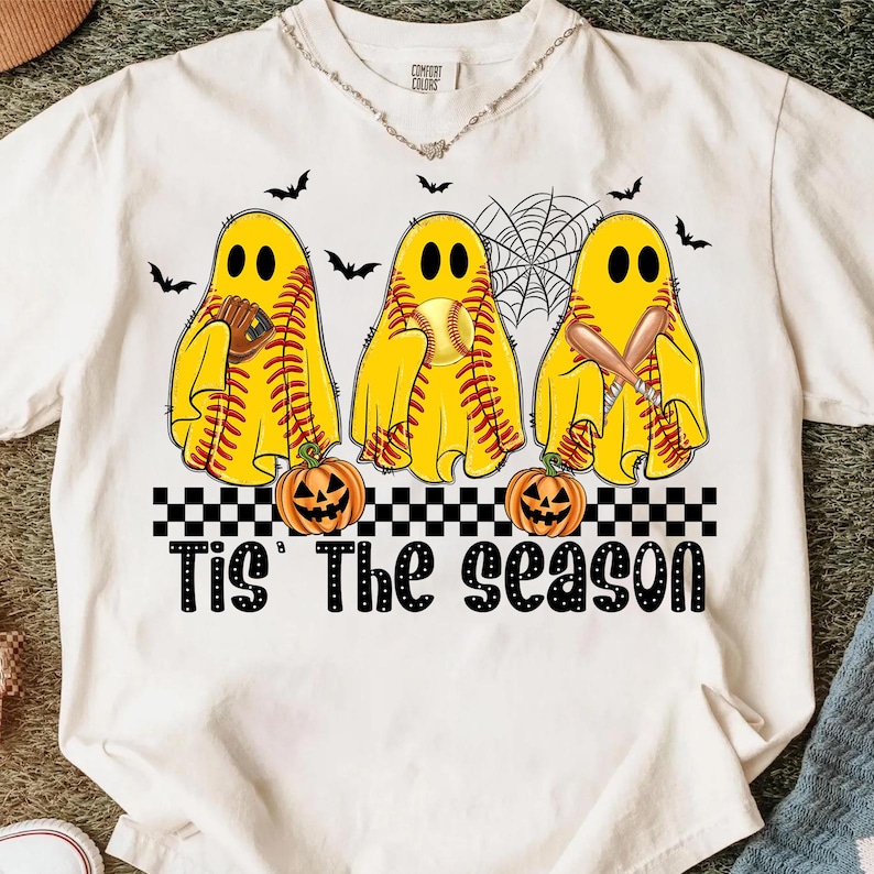 Puede incluir: Camiseta blanca con un dise&ntilde;o de Halloween. Tres figuras de fantasmas amarillos con costuras de b&eacute;isbol sostienen cada una un objeto de b&eacute;isbol. El texto "Tis' The Season" est&aacute; debajo, con calabazas talladas. Tambi&eacute;n hay murci&eacute;lagos y una telara&ntilde;a.