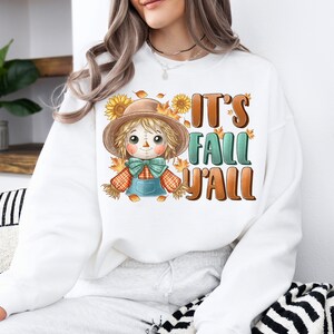 Op de afbeelding: Witte trui met een cartoon-vogelverschrikker en de tekst "It's Fall Y'all".