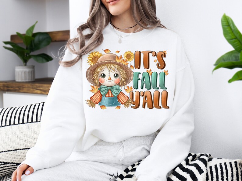 Op de afbeelding: Witte trui met een cartoon-vogelverschrikker en de tekst "It's Fall Y'all".
