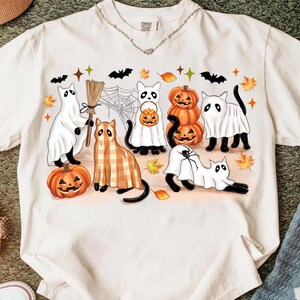 May include: Off-white t-shirt with a Halloween graphic. The design features cartoon cats in ghost costumes, pumpkins, bats, and autumn leaves. The cats are in different poses, with a broom and a spider web.