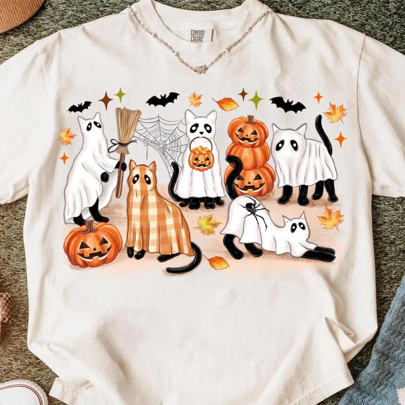May include: Off-white t-shirt with a Halloween graphic. The design features cartoon cats in ghost costumes, pumpkins, bats, and autumn leaves. The cats are in different poses, with a broom and a spider web.
