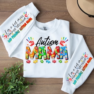 Op de afbeelding: Witte trui met de woorden "Autism Mama" in regenboogkleuren gemaakt van puzzelstukjes en bloemen. De mouwen hebben de tekst "I'm a hot mess always stressed and proudly Autism Mama" in regenboogkleuren.