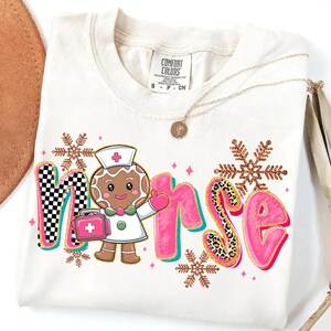 May include: Cream-colored t-shirt with the word "Nurse" in a playful design. The "N" has a black and white checkered pattern, and the "S" has a leopard print. A gingerbread nurse and snowflakes complete the design.