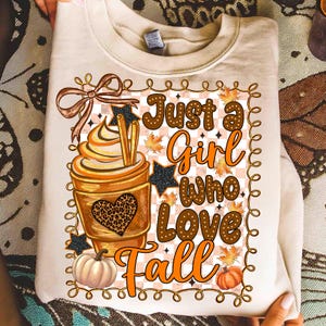 May include: Cream-colored sweatshirt with a fall-themed graphic. The design features a pumpkin spice latte, pumpkins, and the text "Just a Girl Who Love Fall". The graphic includes a bow, stars, and fall leaves.