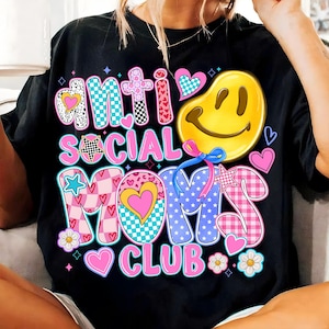 May include: Black t-shirt with a colorful graphic design that says "Anti Social Moms Club". The graphic features a smiley face, stars, hearts, and checkered patterns.