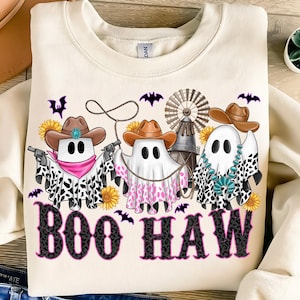 May include: Cream-colored sweatshirt with a graphic of three ghost figures wearing cowboy hats and bandanas, holding a lasso and guns. The text "BOO HAW" is printed in a black and pink leopard print font, with bats and sunflowers.