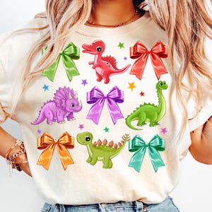 May include: A white t-shirt with a colorful dinosaur and bow design. The dinosaurs are green, purple, and red. The bows are in various colors, including purple, orange, red, and blue.