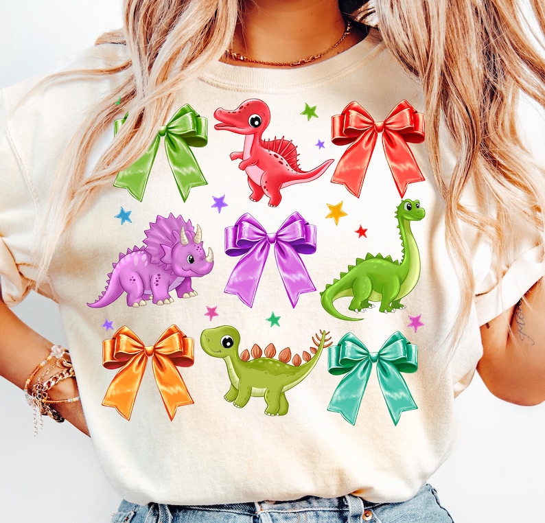 May include: A white t-shirt with a colorful dinosaur and bow design. The dinosaurs are green, purple, and red. The bows are in various colors, including purple, orange, red, and blue.
