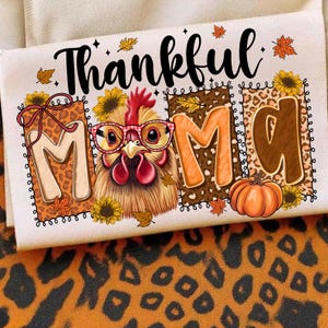 May include: A white t-shirt with the words "Thankful Mom" in black script. The "M" letters have a leopard print design. A cartoon chicken wearing glasses is in the center. A small orange pumpkin is at the bottom.