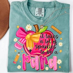 May include: A teal Comfort Colors t-shirt featuring a graphic of a pink apple with a yellow pencil, pink bow, and the text "it takes a lot of sparkles to be a para". The design includes daisies and stars.