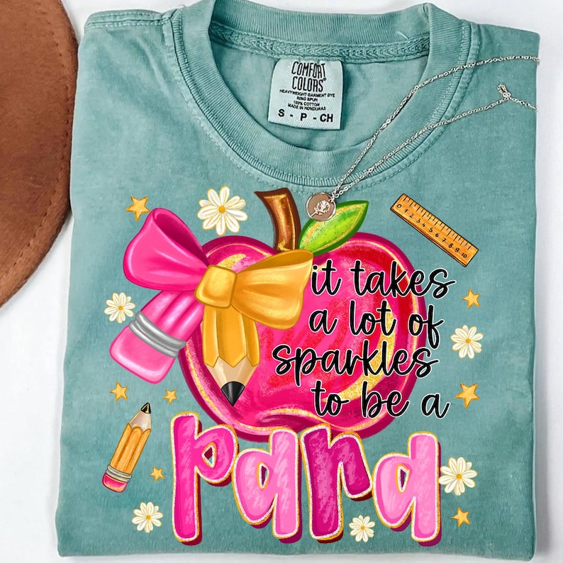 May include: A teal Comfort Colors t-shirt featuring a graphic of a pink apple with a yellow pencil, pink bow, and the text "it takes a lot of sparkles to be a para". The design includes daisies and stars.