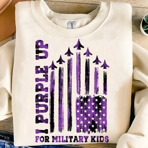 May include: A white sweatshirt with a purple and black design. The design features a purple American flag with a purple star pattern. The text "I Purple Up For Military Kids" is printed on the sweatshirt.